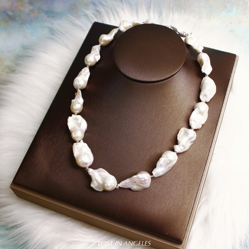 May include: A white baroque pearl necklace displayed on a brown jewelry stand. The pearls are irregularly shaped and strung together with a silver clasp. The necklace is presented on a brown display box with the text "LOST IN ANGELS" at the bottom.