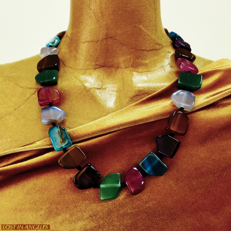 May include: A multi-coloured gemstone necklace featuring polished stones in shades of blue, green, red, brown, and clear. The necklace is strung on a black cord and displayed on a gold-coloured surface. The text "LOST IN ANGELES" is visible at the bottom.