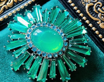 Peut inclure: Une broche vert émeraude vibrante avec une pierre centrale ovale entourée de petites pierres bleues. La broche présente un motif en forme d'éclat de soleil avec des cristaux verts rectangulaires allongés. Le texte "LOST IN ANGELES" est visible en bas.