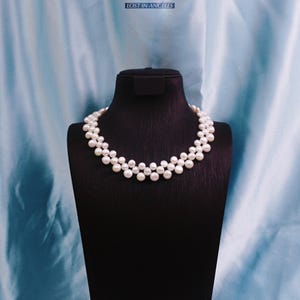 Può includere: Una collana di perle a due fili esposta su un manichino nero. La collana presenta numerose perle bianche, disposte in un design a cascata. Lo sfondo è un tessuto azzurro e il testo "LOST IN ANGELES" è visibile in alto.