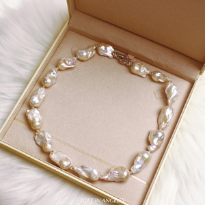 May include: A necklace of large, irregularly shaped, off-white pearls. The pearls are strung together with a delicate gold chain and clasp. The necklace is presented in a beige jewelry box with a gold trim. The text "LOST IN ANGLES" is at the bottom.