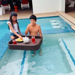 May include: Two children in a swimming pool with a woven tray of food and drinks. The tray holds a plate of food, a decorative white object, and two glasses of red liquid. The pool has blue tile accents.