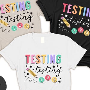 May include: Three t-shirts with the text "TESTING testing 123" in rainbow colors with a pencil and stars. The shirts are in beige, black, and white.