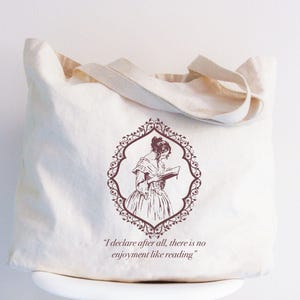 May include: A white canvas tote bag with a burgundy illustration of a woman reading a book inside a decorative frame. The text "I declare after all, there is no enjoyment like reading" is printed below the illustration.