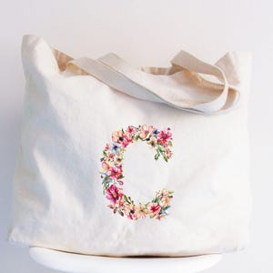May include: Cream-colored tote bag with a floral letter "C" design. The letter is composed of pink, purple, and yellow flowers. The bag has two beige handles and is made of a soft fabric.