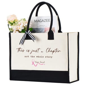 May include: A white canvas tote bag with black trim and handles. The bag has a pink ribbon and the text "This is just a chapter not the whole story Team Sarah" printed on the front.