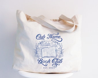 May include: Cream-colored tote bag with blue text and a book-themed design. The text reads "Club Name Book Club EST. 2025." The design features an open book surrounded by stacks of books and foliage. The bag has two handles.