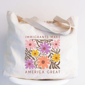 May include: A white canvas tote bag with the text "Immigrants Make America Great" printed in brown. The text is surrounded by a colorful floral design with pink, purple, orange, and yellow flowers.