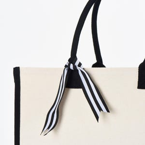May include: A white canvas tote bag with black trim and black handles. The bag has a black and white striped ribbon tied around the handles.