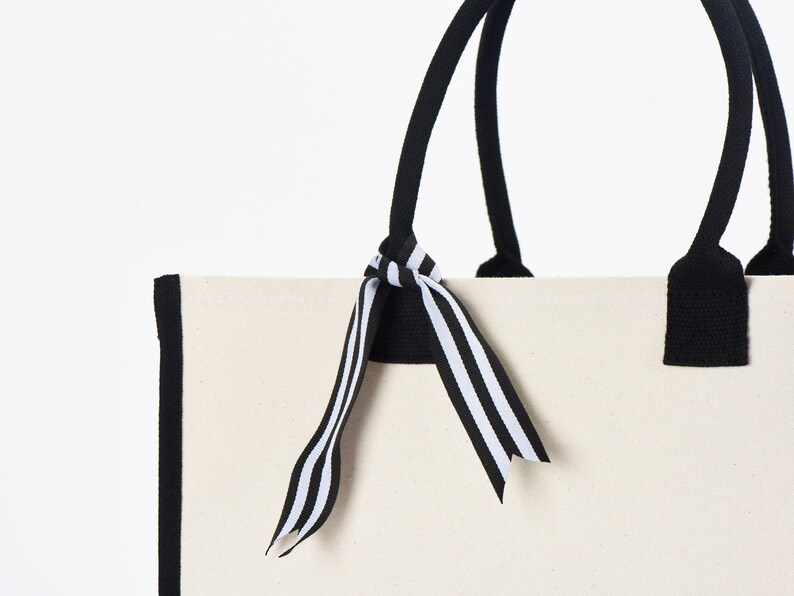 May include: A white canvas tote bag with black trim and black handles. The bag has a black and white striped ribbon tied around the handles.