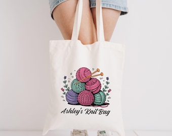 May include: Cream-colored tote bag featuring a colorful illustration of yarn balls and knitting needles. The design includes pink, green, and purple yarn, with the text "Ashley's Knit Bag" in a script font.