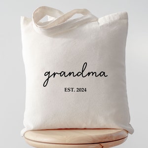 Op de afbeelding: Een witte canvas tote bag met zwarte tekst die "grandma EST. 2024" zegt.