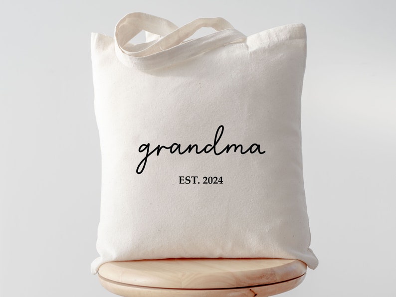 Op de afbeelding: Een witte canvas tote bag met zwarte tekst die "grandma EST. 2024" zegt.