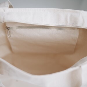 May include: A white canvas tote bag with a zippered pocket on the inside. The bag is empty and the zipper is closed.