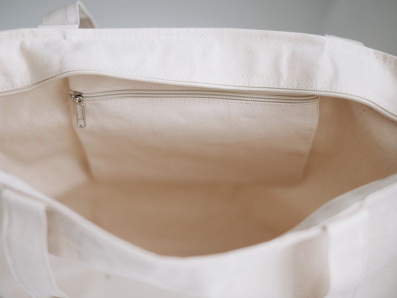 May include: A white canvas tote bag with a zippered pocket on the inside. The bag is empty and the zipper is closed.