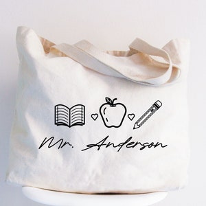 May include: A white canvas tote bag with black text and illustrations. The text reads "Mr. Anderson" and the illustrations are an open book, an apple, two hearts, and a pencil.