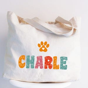 May include: A white canvas tote bag with a paw print and the name "CHARLIE" printed in colorful letters. The letters are filled with a pattern of paw prints and bones.
