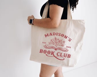 May include: Cream-colored tote bag with a red design featuring the text "Madison's Book Club" and an illustration of an open book with flowers. The year "2025" is also included in the design. The bag is carried by a person.