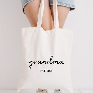 Op de afbeelding: Een witte canvas tote bag met de tekst "grandma EST. 2024" in zwart gedrukt.