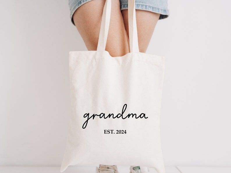 Op de afbeelding: Een witte canvas tote bag met de tekst "grandma EST. 2024" in zwart gedrukt.