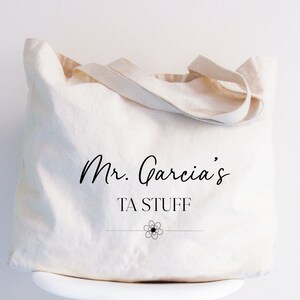 May include: A white canvas tote bag with black text that reads "Mr. Garcia's TA STUFF" with a small flower design underneath.
