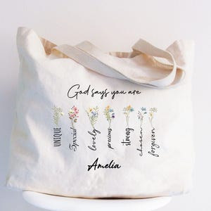 May include: A white canvas tote bag with the text "God says you are" and a floral design. The words "unique, special, lovely, precious, strong, chosen, forgiven" are written vertically below the flowers. The name "Amelia" is written at the bottom of the bag.