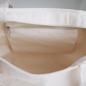 May include: Cream-colored canvas tote bag with a zippered top and an interior pocket. The bag has sturdy handles and a simple, minimalist design. The zipper and pocket are a light silver color.