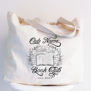 May include: A white canvas tote bag with black text and a graphic of a stack of books with a single open book on top. The text reads "Club Name Book Club Est. 2024".