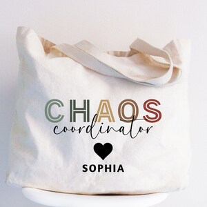 May include: Cream-colored canvas tote bag with the words "CHAOS coordinator" in a mix of colors and fonts. A black heart and the name "SOPHIA" are below the text. The bag has two handles.