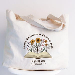 May include: Cream-colored tote bag with a floral design and text. The design features an open book with colorful flowers and the text "Escuela del Servicio de Precursor 2025" and "LA MEJOR VIDA Amelia". The bag has matching cream-colored handles.