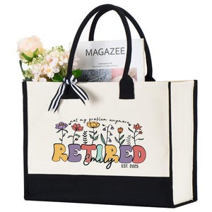 May include: Cream-colored tote bag with black accents and handles. The bag features the text "Retired Emily Est. 2025" in colorful letters with floral accents. The bag contains flowers and a magazine.