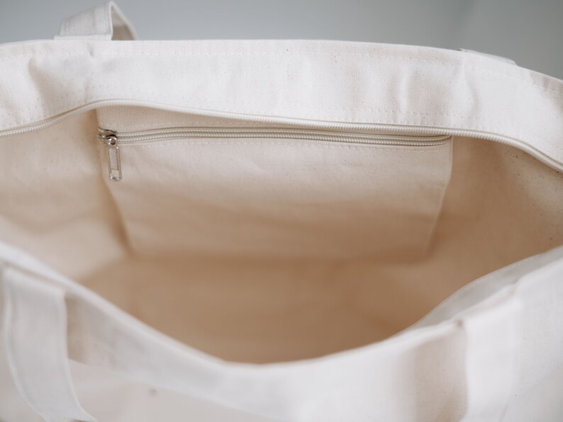 Op de afbeelding: Een witte canvas tote bag met een ritsvak aan de binnenkant. De tas is leeg en de rits is dicht.