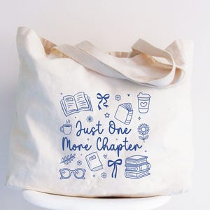May include: Cream-colored tote bag with blue line art of books, coffee cups, bows, and flowers. The text "Just One More Chapter" is written in a script font. The bag has a rectangular shape with two handles.