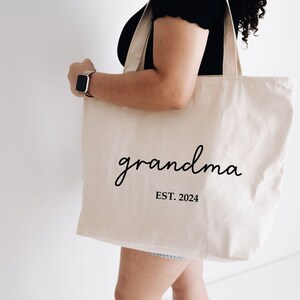 Op de afbeelding: Een witte canvas tote bag met zwarte tekst die "grandma EST. 2024" zegt.