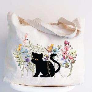 May include: A cream-colored tote bag featuring a black cat silhouette amidst a colorful floral design. The flowers include pink, blue, yellow, and purple blooms, with a dragonfly above. The bag has two beige handles.