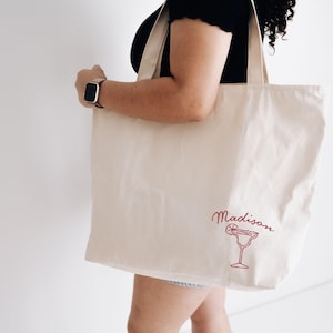 May include: A beige canvas tote bag with the name "Madison" in cursive script and a red margarita glass illustration. The bag has long straps and is held by a person. The background is a plain white wall.