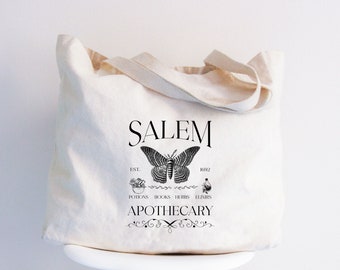 Könnte beinhalten: Cremefarbene Tragetasche mit schwarzem Grafikdesign. Das Design enthält das Wort "SALEM" über einer detaillierten Schmetterlingsillustration. Darunter stehen die Wörter "POTIONS BOOKS HERBS ELIXIRS" und "APOTHECARY".