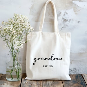 Op de afbeelding: Witte canvas tote bag met zwarte tekst die "grandma est. 2024" zegt. De tas ligt op een houten oppervlak met een boeket witte bloemen in een glazen vaas.