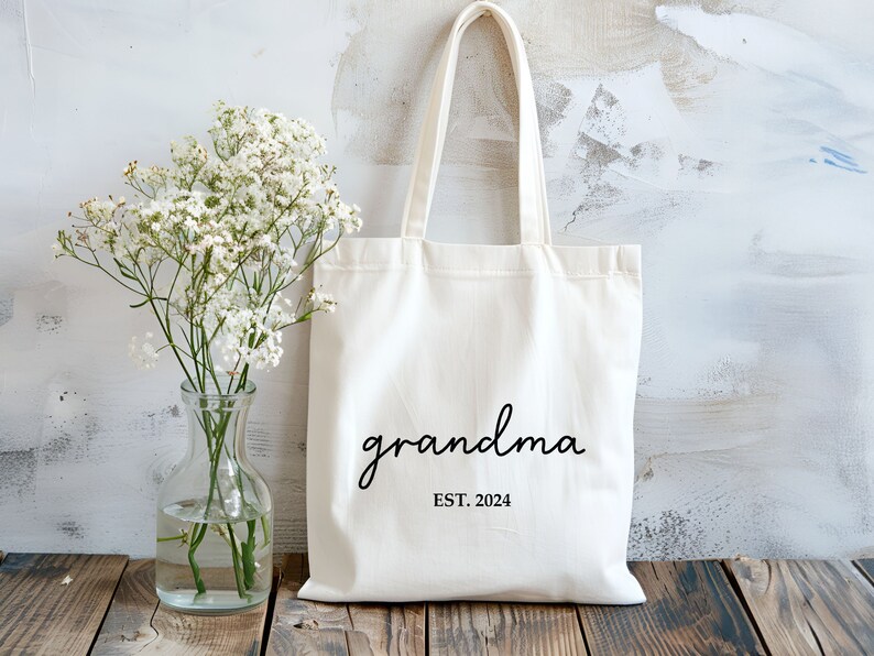 Op de afbeelding: Witte canvas tote bag met zwarte tekst die "grandma est. 2024" zegt. De tas ligt op een houten oppervlak met een boeket witte bloemen in een glazen vaas.