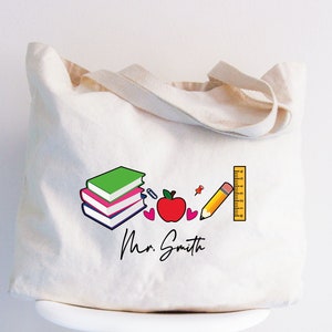 May include: A white canvas tote bag with a colorful design featuring books, an apple, a pencil, a ruler, and two hearts. The text "Mr. Smith" is printed in black cursive.