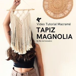 Puede incluir: Un tapiz de macramé blanco con una barra de madera en la parte superior. El tapiz tiene la forma de una V grande con flecos que cuelgan de la parte inferior. El texto "Video Tutorial Macrame TAPIZ MAGNOLIA By Mar de Comodoro" es visible en la imagen.