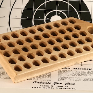 Peut inclure: Un support de munitions en bois clair avec de nombreux trous circulaires. Le support a une poignée arrondie et est posé contre une cible et des instructions imprimées. Le grain du bois est visible.