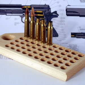 May include: A wooden ammunition loading block with multiple holes, holding several brass bullet casings and live rounds. The block is made of light-colored wood and is set against a backdrop of technical diagrams.