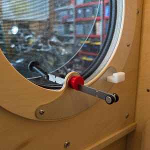 May include: Close-up of a wooden window frame with a round window and a black mesh screen. The window has a red handle and a metal arm for opening and closing. The frame is light-colored wood.