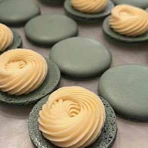 Puede incluir: Primer plano de varios macarons verde pastel, algunos cubiertos con remolinos de crema de mantequilla amarillo pálido. Los macarons están dispuestos sobre una superficie clara, mostrando los delicados detalles de los productos horneados.
