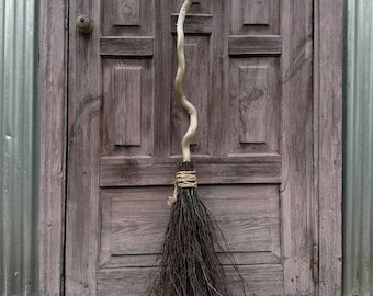 May include: A broom featuring a long, light-colored, winding wooden handle that splits into a Y-shape at the top. The base consists of dark, thin, natural twigs bound together with a thick, light-colored twine cord wrapped around the top.