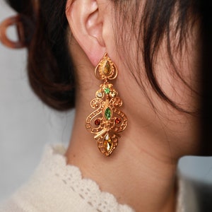 May include: Gold dangle earrings with a filigree design and colorful gemstones. The earrings feature green, red, and yellow stones.