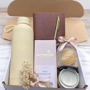 May include: A gift box filled with items including a yellow water bottle, a brown leather journal with a gold pen, a pink and white chocolate bar, a small burlap bag with a pink ribbon and a label that says "Caramels", and a white candle in a glass jar.