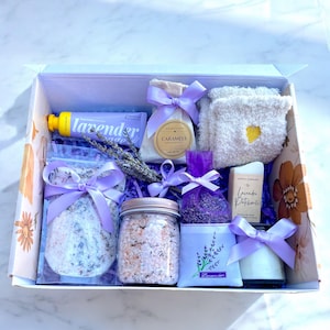 May include: A lavender-themed gift box filled with bath products, including bath salts, a candle, a hand cream, and a lavender sachet. The box is lined with purple tissue paper and decorated with lavender flowers.