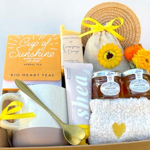 May include: A gift basket with a yellow and white theme. It includes a mug, tea, honey, shea butter, a washcloth, and a small bag with a sunflower.